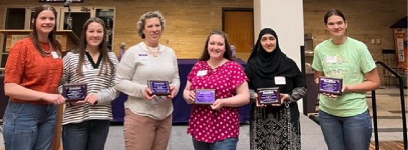 Six people standing in front of a stage, each holding a purple plaque. 