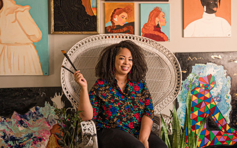 A black woman is sitting in a white wicker chair, holding two paintbrushes in her right hand. She is wearing a black floral short-sleeve top and black pants. She is sitting in front of various pieces of art on the back wall. 